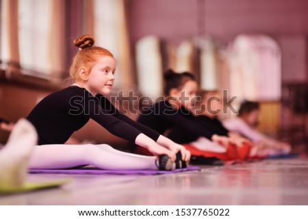 child preschooler - a cute red-haired girl ballerina stretching and doing the splits on the background of ballet schools and groups of children