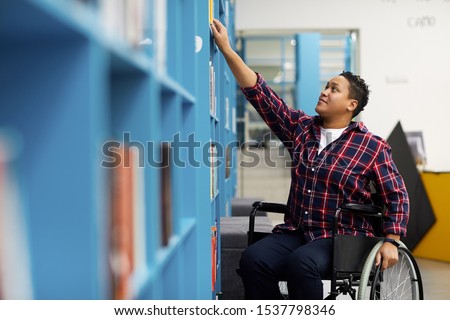 Portrait of disabled student in wheelchair choosing books while studying in college library, copy space Royalty-Free Stock Photo #1537798346