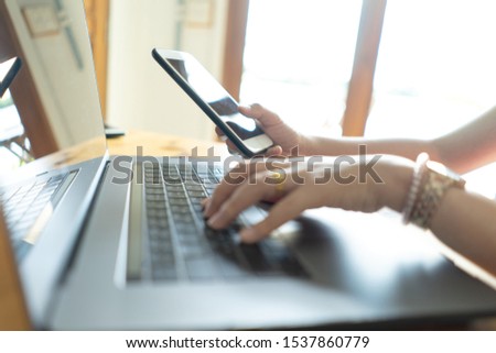 Young business woman holding mobile phone and typing on laptop computer keyboard in coffee shop.
Female freelance working on notebook computer in cafe.