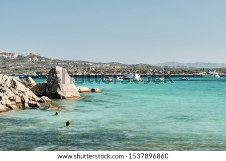 A beautiful shot of rocks in the water with boats and mountains in the distance under a blue sky