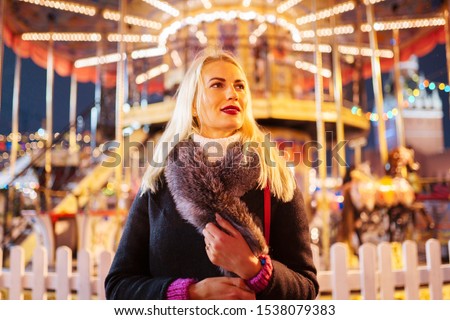 Picture of young blonde woman in coat with fur collar in park on background of carousel