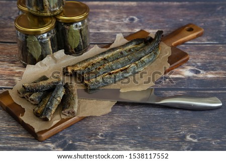 Fried lamprey, on a wooden board with baking paper. A knife for slicing it and glass jars of fish. Stage of conservation. Royalty-Free Stock Photo #1538117552
