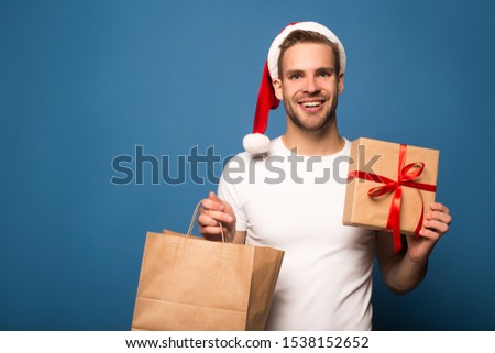 smiling young man in santa hat holding paper bag and present isolated on blue