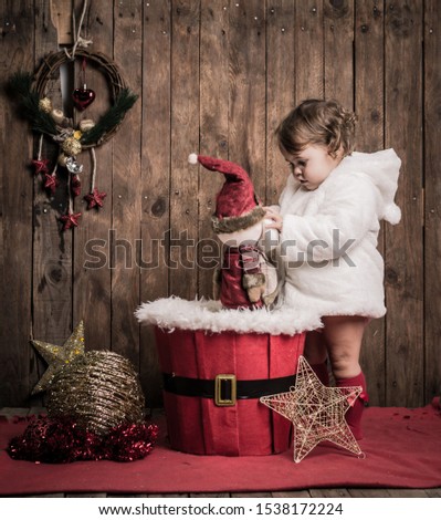 Cute child in Christmas time. Funny kid playing with a toy on Christmas stage.  Little blonde girl is wearing a white coat on a wooden background. Xmas winter holiday concept.