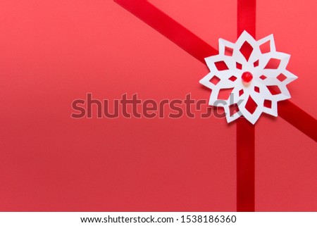 Single hand cut white paper snowflake laying on red ribbon ribbon on red festive background. Photo with blank space.