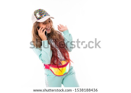 Teen girl with long blonde hair dyed with tips pink, in shiny white cap, light blue sports suit, belt bag smiles and speaks on phone