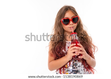 Teen girl with long blonde hair dyed with tips pink, in shiny light dress, black and white sneakers, glasses, standing with headphones, holding juice in glass bottle with striped tube in hand