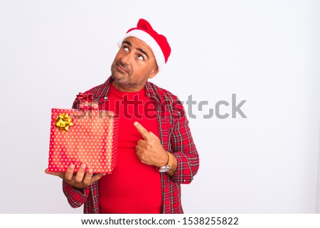 Middle age man wearing Christmas Santa hat holding gift over isolated white background serious face thinking about question, very confused idea