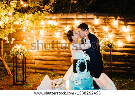 Bride and a groom is kissing near rustic wedding cake outdoors. Cake with delicate blue and white flowers. Couple in magical evening forest decorated light garlands. Night ceremony.