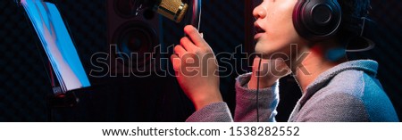 Asian Teenager man black hair earmuff headphone sing a song loudly power sound over hanging microphone condenser, speakers, composer note book and monitor. Studio Sound Proof Absorbing wall room Royalty-Free Stock Photo #1538282552
