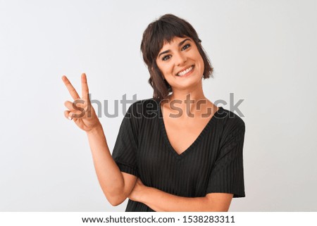 Young beautiful woman wearing black t-shirt standing over isolated white background smiling with happy face winking at the camera doing victory sign. Number two.