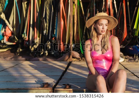 Image of serious caucasian woman in swimsuit and straw hat looking aside while sitting near surfboards and sails on beach
