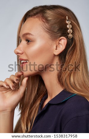  Profile portrait shot of a lady with blond hair, posing on a blue background. She has a  hair clip, decorated with four golden  coins. The girl is wearing a dark blue T-shirt Royalty-Free Stock Photo #1538317958