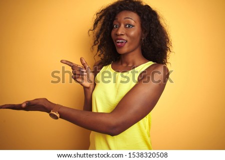 Young african american woman wearing t-shirt standing over isolated yellow background amazed and smiling to the camera while presenting with hand and pointing with finger.