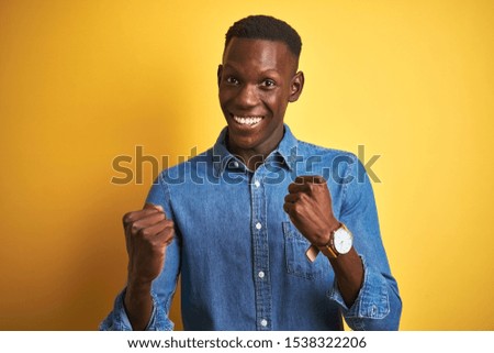 Young african american man wearing denim shirt standing over isolated yellow background celebrating surprised and amazed for success with arms raised and open eyes. Winner concept.