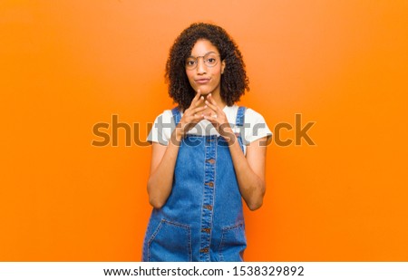young pretty black woman scheming and conspiring, thinking devious tricks and cheats, cunning and betraying against orange wall