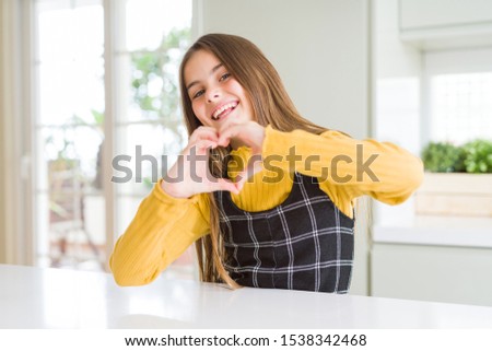 Young beautiful blonde kid girl wearing casual yellow sweater at home smiling in love doing heart symbol shape with hands. Romantic concept.
