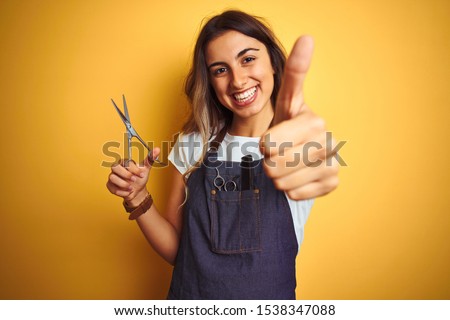 Young beautiful hairdresser woman holding scissors over yellow isolated background happy with big smile doing ok sign, thumb up with fingers, excellent sign