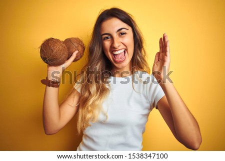 Young beautiful woman holding coconut over yellow isolated background very happy and excited, winner expression celebrating victory screaming with big smile and raised hands