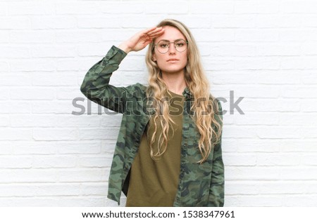 young blonde woman greeting the camera with a military salute in an act of honor and patriotism, showing respect against brick wall background