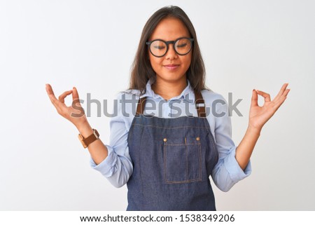 Young beautiful chinese woman wearing glasses and apron over isolated white background relax and smiling with eyes closed doing meditation gesture with fingers. Yoga concept.