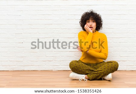 young pretty afro woman open-mouthed in shock and disbelief, with hand on cheek and arm crossed, feeling stupefied and amazed sitting on the floor