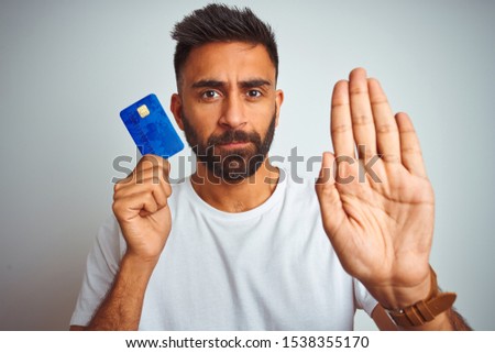Young indian customer man holding credit card standing over isolated white background with open hand doing stop sign with serious and confident expression, defense gesture