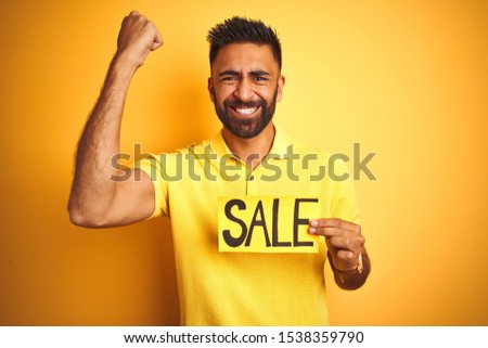 Young arab indian hispanic man holding sale banner over isolated yellow background annoyed and frustrated shouting with anger, crazy and yelling with raised hand, anger concept