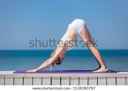 Sporty young woman standing in Downward facing dog exercise, adho mukha svanasana pose. Slim girl practicing yoga outdoor by sea, blue sky, wooden terrace. Calm, relax, healthy lifestyle concept