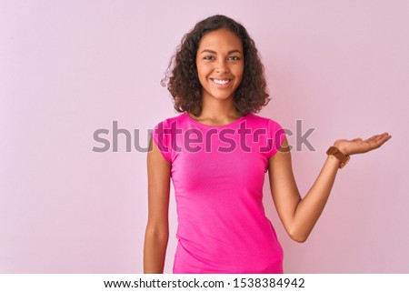 Young brazilian woman wearing t-shirt standing over isolated pink background smiling cheerful presenting and pointing with palm of hand looking at the camera.