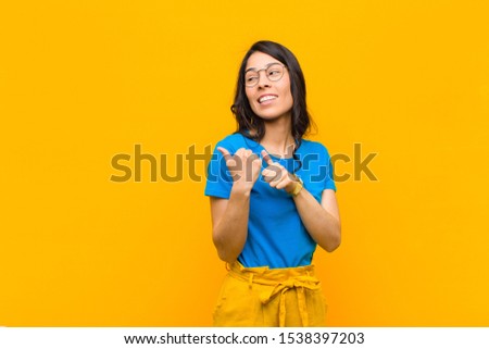 young pretty latin woman smiling cheerfully and casually pointing to copy space on the side, feeling happy and satisfied against orange wall