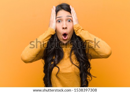 young pretty hispanic woman looking unpleasantly shocked, scared or worried, mouth wide open and covering both ears with hands against brown wall