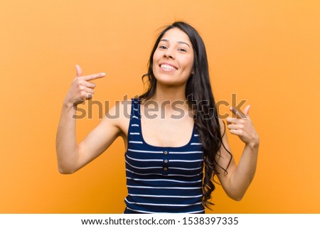 young pretty hispanic woman smiling confidently pointing to own broad smile, positive, relaxed, satisfied attitude against brown wall