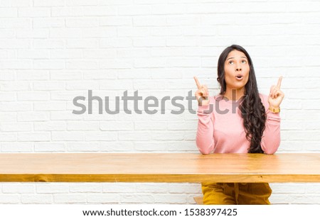 young  pretty latin woman feeling awed and open mouthed pointing upwards with a shocked and surprised look sitting in front of a table
