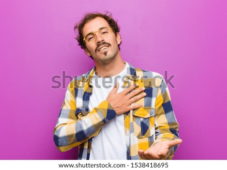 young handsome man feeling happy and in love, smiling with one hand next to heart and the other stretched up front against purple wall
