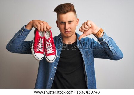 Young handsome man holding casual sneakers standing over isolated white background with angry face, negative sign showing dislike with thumbs down, rejection concept