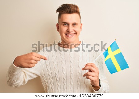 Young handsome man holding Sweadish Sweden flag over isolated white background with surprise face pointing finger to himself