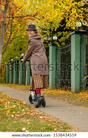 Beautiful girl in hat and glasses riding a scooter on a background of autumn. Modern young woman resting in the park or going to work.