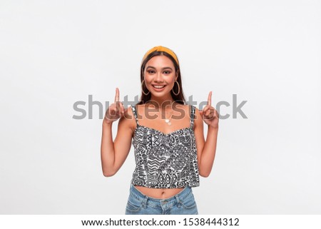 Pretty african-american teenager in a headband, sleeveless t-shirt and jeans pointing at the empty space in a upper side of a frame standing isolated over white background. Copy space of advertising.