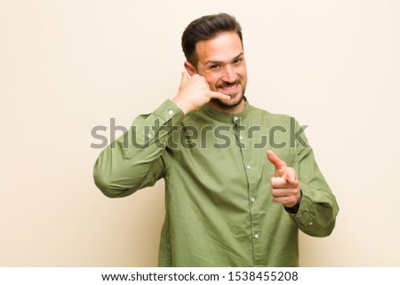 young handsome man smiling cheerfully and pointing to camera while making a call you later gesture, talking on phone