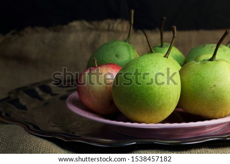 Green organic pears and one small red apple, different not like others, on delicate pink plate on silver metal tray, picturesque still life on coarse retro cloth canvas. Dark atmospheric fall harvest