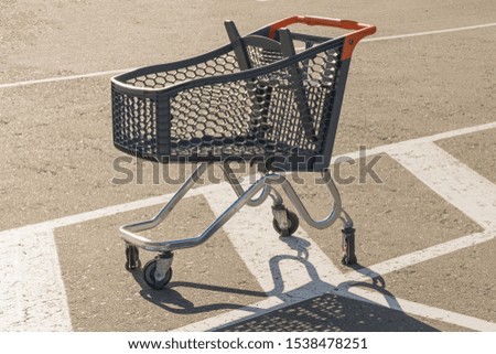 A metal shopping trolley on wheels stands in a parking lot in front of a supermarket on asphalt with white markings in the sun and cast a shadow on the asphalt