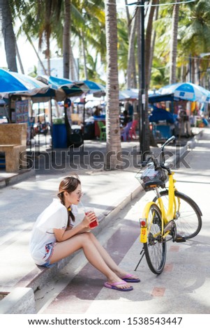 Cute asian woman sitting on the beach street drinking soft drink from plastic cup beside yellow bike.