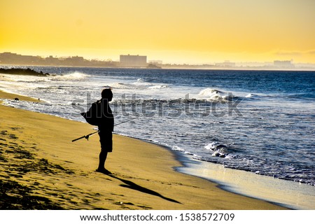 silhouette of a fisherman wearing a backpack holding a fishing pole on the beach shore looking at the ocean water with waves and cityscape in the background