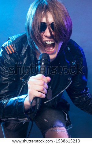 The young guy in black leather jacket is singing in microphone rock music on blue background.