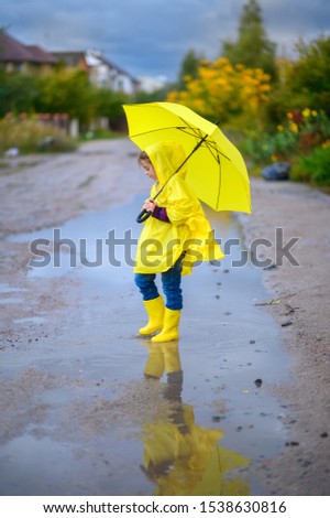 Cute little girl in a raincoat and with a yellow umbrella in her hands walks through puddles, walks in boots and squirts
