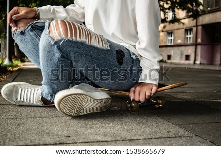 A girl sitting on a skateboard. White sweater end sport shoe and blue jeans pants.