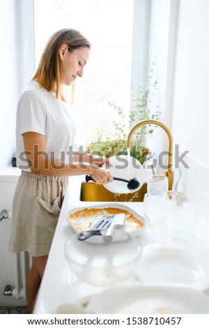 Beautiful smiling young woman washing the dishes in modern white kitchen.