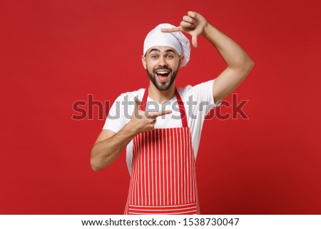 Excited young bearded male chef cook or baker man in striped apron toque chefs hat posing isolated on bright red background. Cooking food concept. Mock up copy space. Making hands photo frame gesture