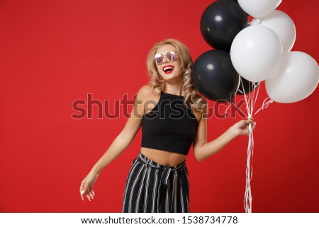 Laughing young woman girl in black clothes, eyeglasses posing isolated on red wall background. St. Valentine's Day birthday holiday party concept. Mock up copy space. Celebrating holding air balloons